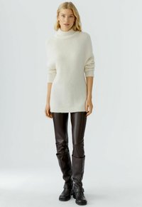 Pull en tricot blanc avec un col montant, coupe oversize et manches trois-quarts, associé à un pantalon en cuir marron ajusté et des bottes marron hautes.