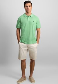 Polo vert avec col, manches courtes et un logo. Associé à un short beige en tissu léger et des chaussures beiges à enfiler.