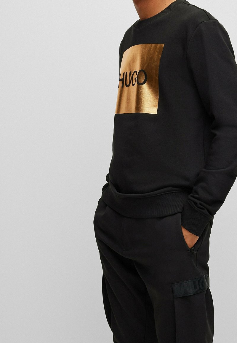 Sweat-shirt noir avec un logo rectangulaire en dorure portant l'inscription « HUGO ». Poignets et ourlet côtelés. Complété par un pantalon de jogging noir.