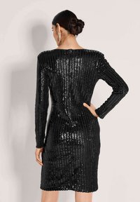 Frau mit einem glatten Dutt, die ein schwarzes, langärmliges, knielanges Kleid mit vertikalen Paillettenstreifen trägt und mit einer Hand auf der Hüfte posiert.