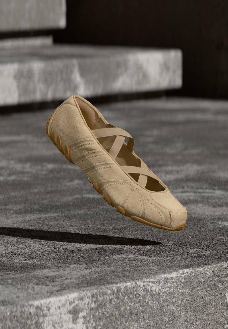 Beige Mary Jane platte schoen met gekruiste elastische bandjes zwevend boven grijze betonnen treden.