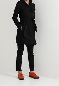 Černý trenčkot s páskem v pase, bočními kapsami a širokým límcem. Kombinován s černými kalhotami a jasně oranžovými botami.