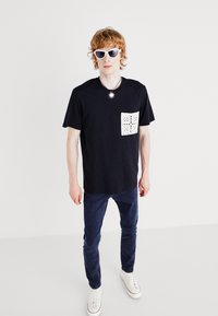 T-shirt en coton noir avec un design de poche perforée blanche, associé à un jean bleu foncé et des baskets blanches, porté avec des lunettes de soleil blanches.