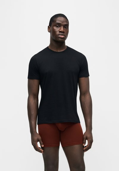 Homme debout portant un t-shirt noir uni à manches courtes et un short ajusté couleur rouille sur un fond clair uni.