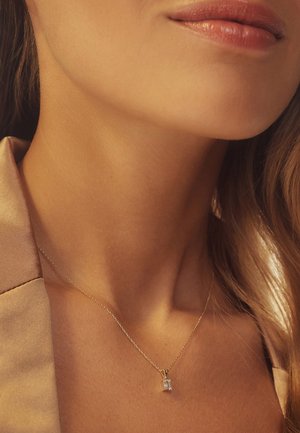 Collier en chaîne dorée avec un pendentif rectangulaire arborant une pierre précieuse claire, posé sur un teint neutre et un fond de tissu beige doux.