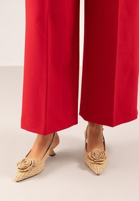 Zapatos slingback de tejido beige con punta afilada, con un acento decorativo en forma de rosa, combinados con pantalones rojos anchos. Tacón bajo en bloque.