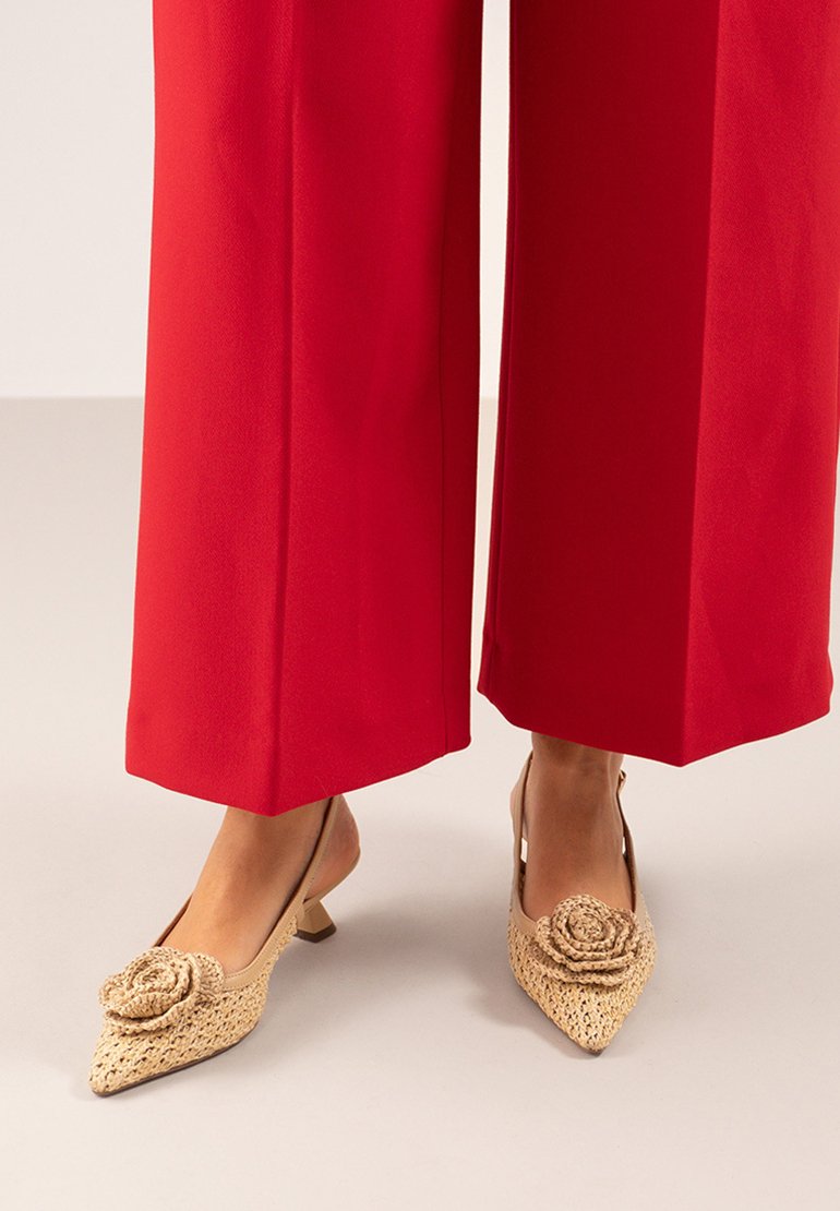 Zapatos slingback de tejido beige con punta afilada, con un acento decorativo en forma de rosa, combinados con pantalones rojos anchos. Tacón bajo en bloque.