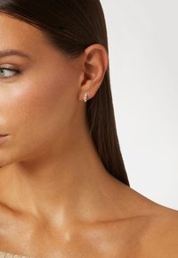 Boucles d'oreilles en argent avec trois pierres précieuses transparentes disposées en ligne, portées sur l'oreille d'un modèle. Fini métallique lisse et design minimaliste.