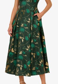 Zelené květinové šaty s plnou sukní, zdobené zlatými akcenty a texturovanou tkaninou. Obsahují kapsy, mají zakázkový design a přiléhavou tělesnou část.