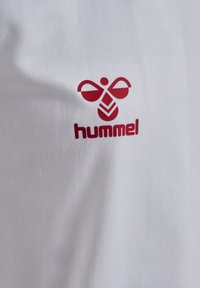 Vit t-shirt med en texturerad yta som har en röd logotyp av en stiliserad bi och varumärket "hummel" under.