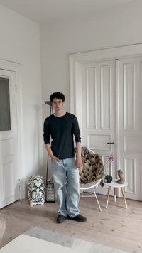 A young person in a ribbed dark top and light blue jeans stands in a room with white walls and wooden floors, next to decorative objects.