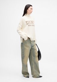 Maglione crema con la scritta "DKNY NEW YORK" in lettere marroni, abbinato a jeans a gamba larga verde chiaro. Accessoriato con una borsa nera.