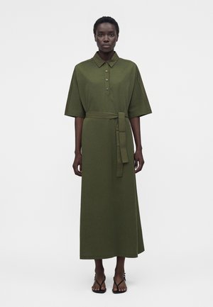 Γυναίκα στέκεται φορώντας μια midi φορεματάκι σε ελαιί πράσινο με κουμπιά, ζώνη, κοντά μανίκια και μαύρα σανδάλια με λουριά.