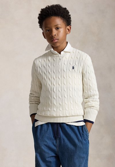 Dreng med hænderne i lommerne iført en hvid kabelstrikket sweater over en hvid skjorte med krave og løse blå jeans mod en neutral baggrund.