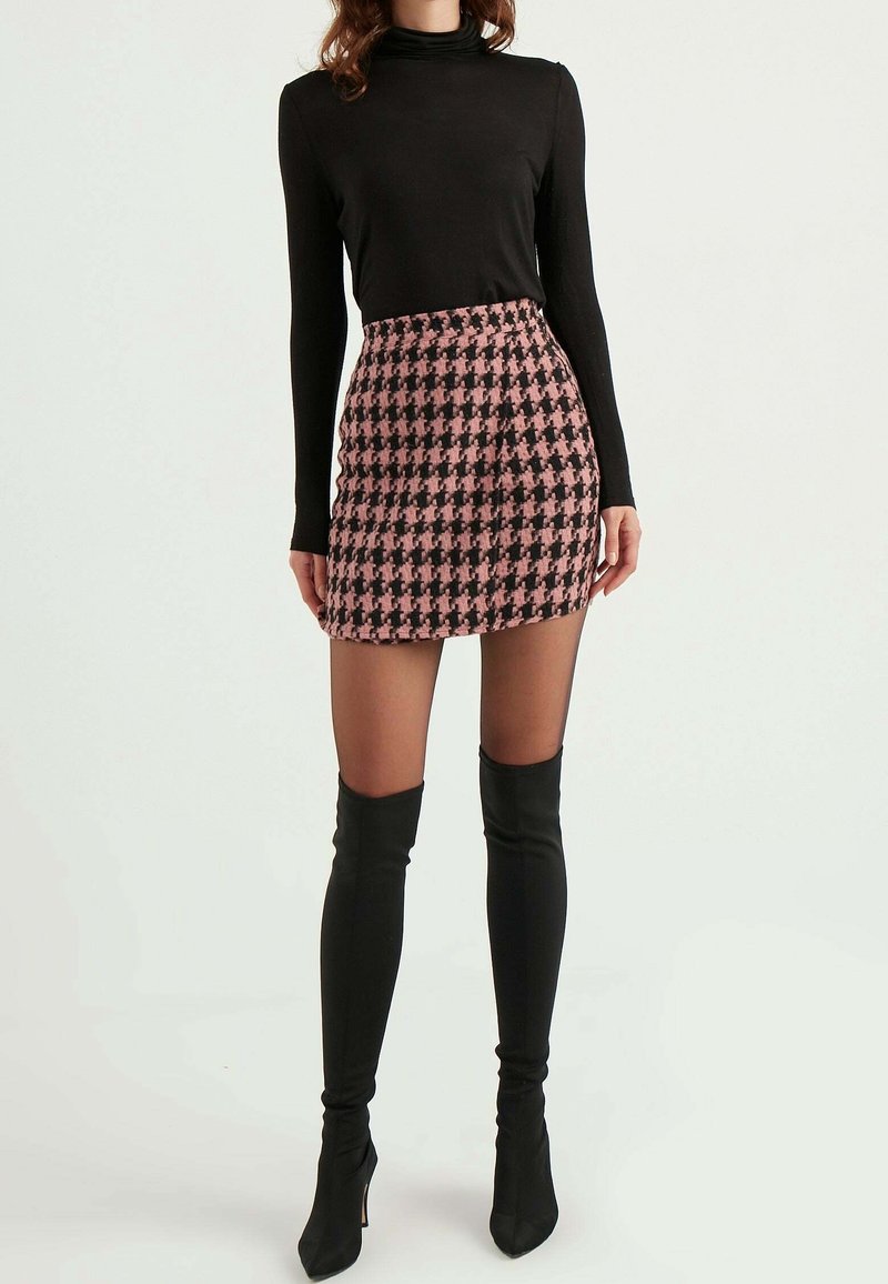 Femme portant un pull à col roulé noir à manches longues, une mini-jupe pied-de-poule rose et noire, des collants transparents et des bottes noires au-dessus du genou, debout.