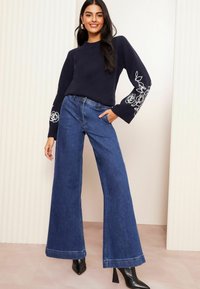 Pulover marin cu broderie florală pe mâneci, asortat cu blugi evazați albaștri și botine negre cu toc. Siluetă curată și modernă.