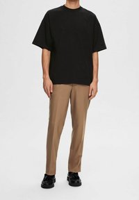 Hombre vestido con una camiseta negra de manga corta y holgada, pantalones beige de pierna recta y mocasines negros, sobre un fondo blanco sencillo.