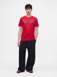 Jeune homme debout portant un t-shirt rouge avec un texte bleu, un pantalon noir ample et des chaussures bordeaux sur un fond blanc uni.