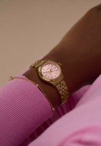 Reloj de pulsera de metal dorado con esfera rosa, bisel texturizado y marcadores de hora dorados, combinado con una delicada pulsera dorada, llevado en una muñeca con un suéter rosa.