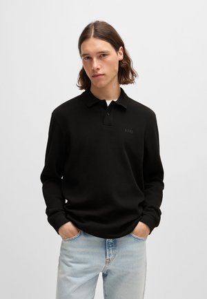 Zwarte polo met lange mouwen, een kraag, enkele knoopsluiting en subtiele branding op de borst, gecombineerd met lichtblauwe jeans.