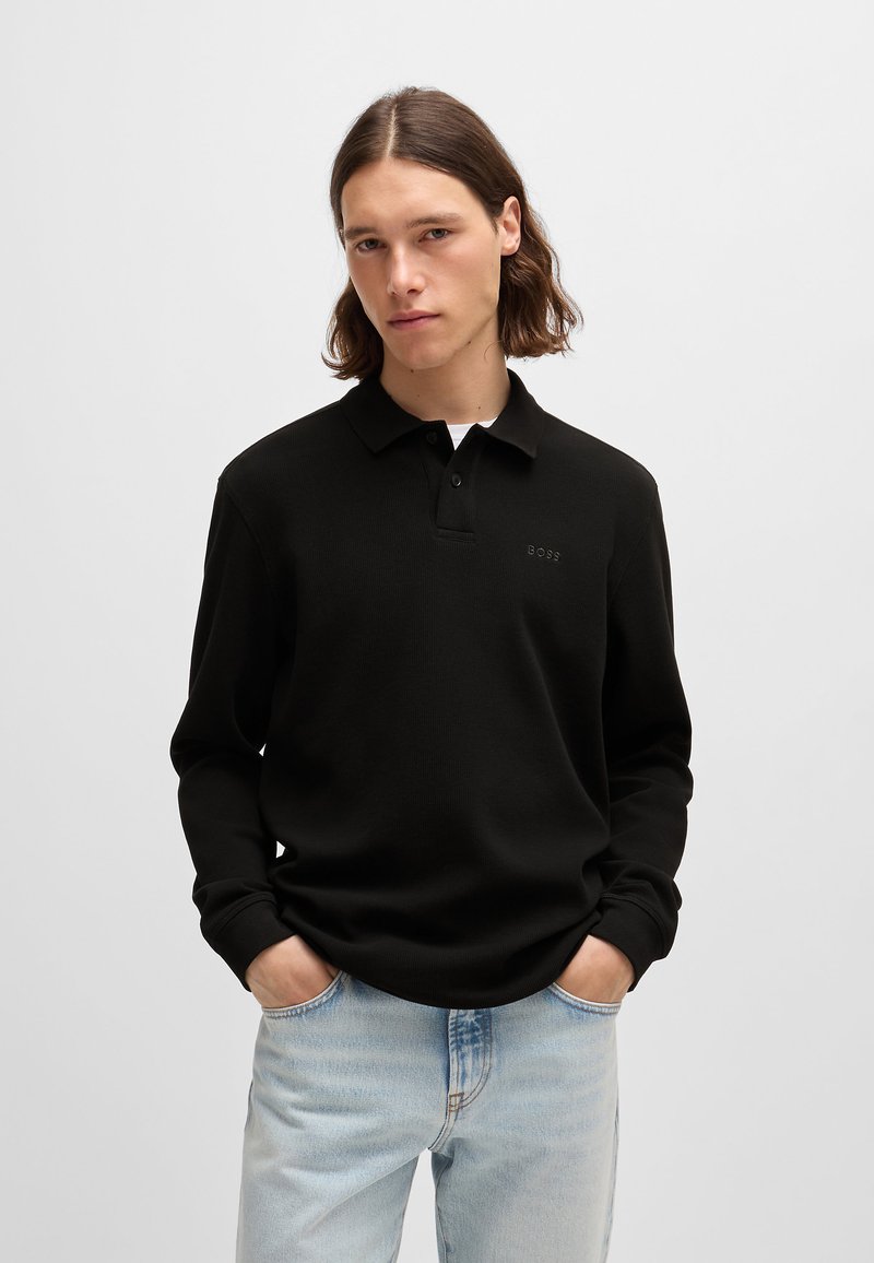 Camisa polo preta de manga longa com colarinho, fecho de botão único e marca discreta no peito, combinada com jeans azul claro.