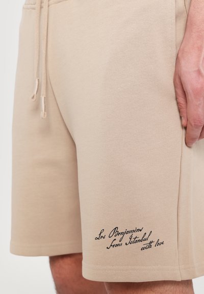 Shorts en coton beige avec une taille élastique, un cordon de serrage et une poche latérale. Présente un texte en cursive sur l'ourlet : "Les Benjamin d'Istanbul avec amour."