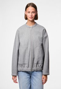 Szara bomberka na zamek z okrągłym dekoltem, dwoma przednimi kieszeniami, elastycznym pasem i miękkim, teksturowanym materiałem. Noszona na niebieskich dżinsach.