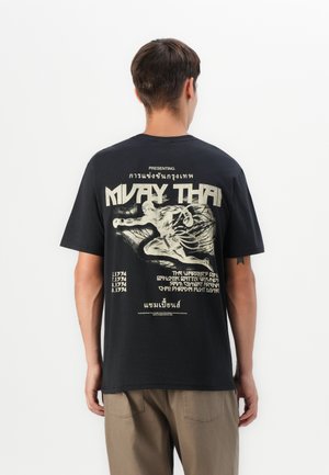 Mann trägt ein schwarzes Muay Thai Grafik-T-Shirt mit einer Illustration eines Boxkämpfers und Text in Englisch und Thai, von hinten betrachtet.