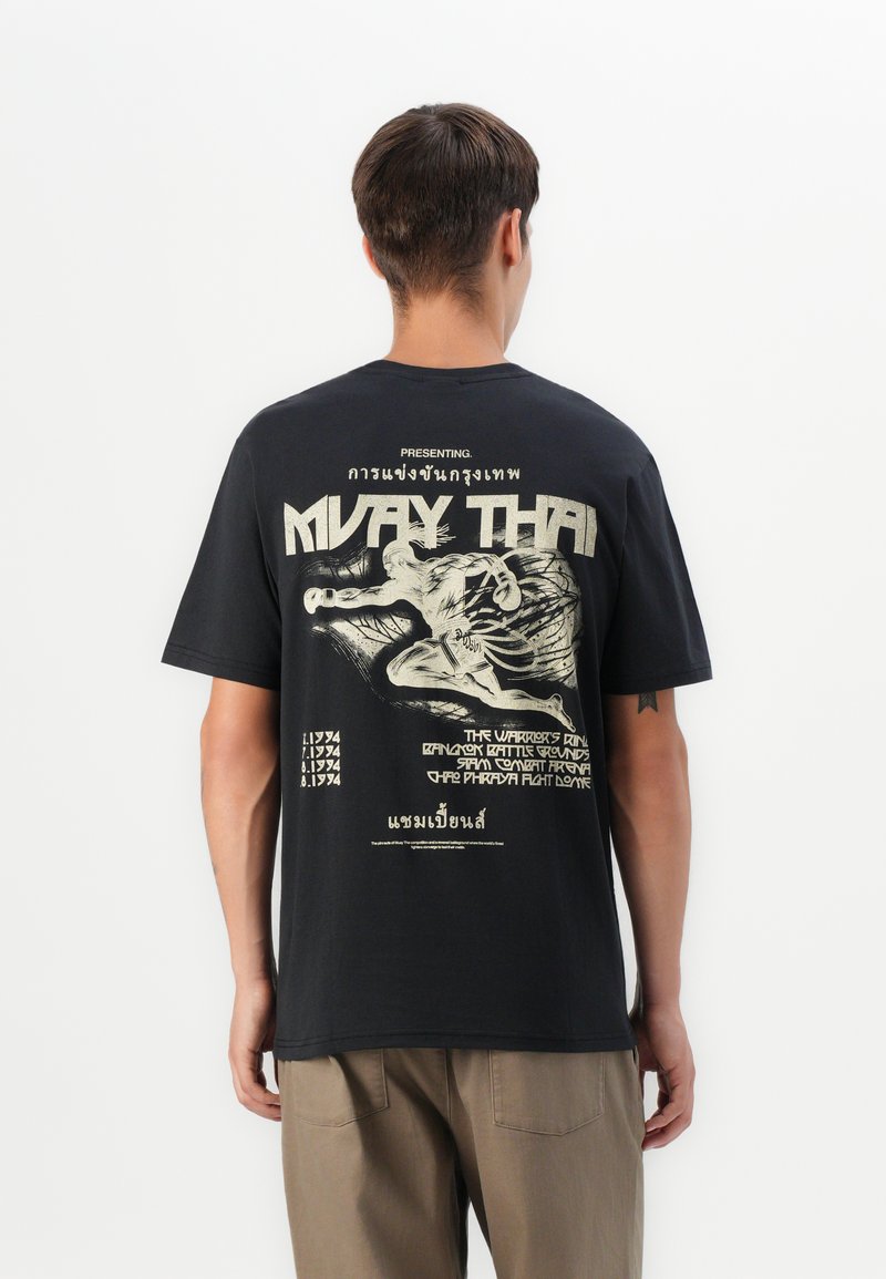 Man wearing black Muay Thai graphic t-shirt with boxing fighter illustration and text in English and Thai, viewed from the back.