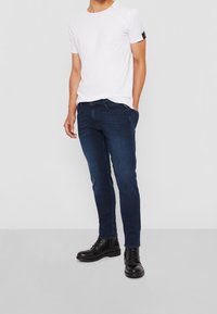 Man som bär mörkblå slim jeans, svarta snörskor och en enfärgad vit t-shirt med kort ärm, stående med en hand i fickan.