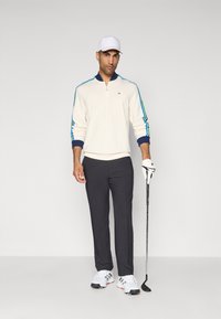 Hvit langermet polo med blå og grønne striper på ermene, mørke bukser, hvit caps, holder en golfklubbe, og iført hvite golfsko.