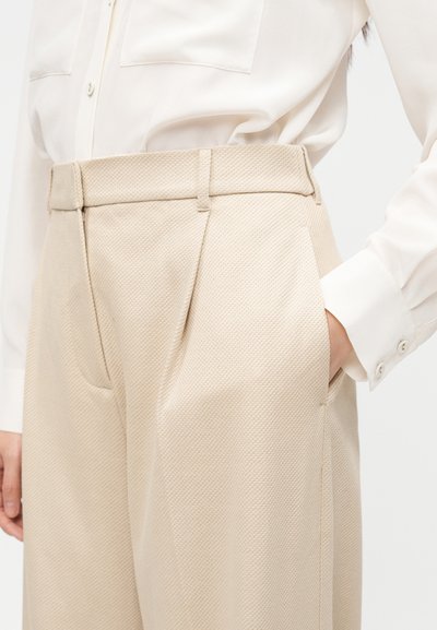 Γυναίκα που φοράει μπεζ παντελόνι με υφή και ψηλή μέση με πλαϊνές τσέπες και λευκό πουκάμισο με κουμπιά και μπροστινές τσέπες στο στήθος.