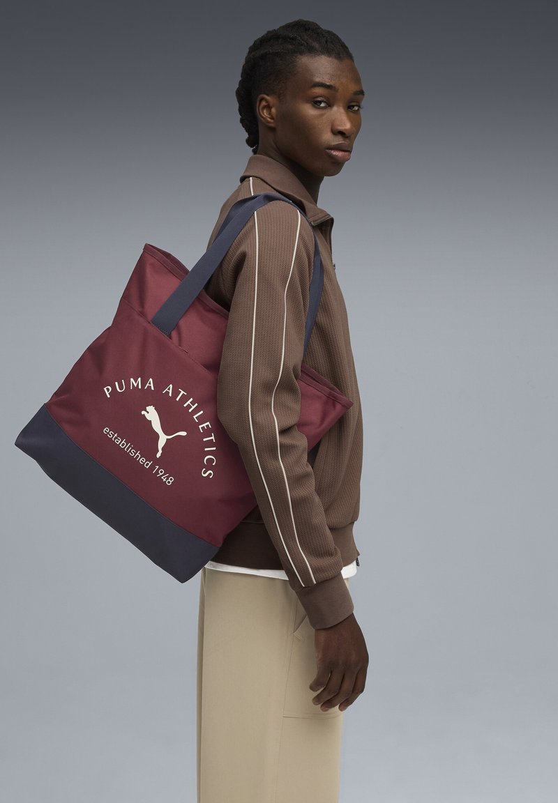 Bordeaux-kangaslaukkui, jossa on sininen pohja, jossa on 'PUMA ATHLETICS' -logo ja perustamisvuosi. Valmistettu kestävästä materiaalista, jossa on kaksi olkahihnaa.