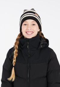 Veste matelassée noire avec une fermeture éclair à l'avant et un col montant, associée à un bonnet en tricot noir et blanc à rayures avec un petit patch en cuir.