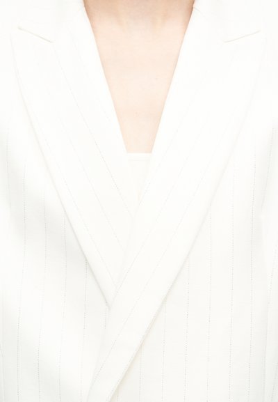 Gros plan sur un torse portant un blazer blanc à fines rayures avec larges revers, par-dessus un haut clair, sur fond uni.