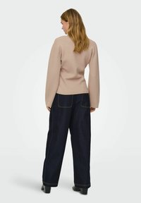 Pull beige clair avec des manches longues et larges, cintré à la taille, associé à un jean large taille haute bleu foncé avec des coutures visibles.