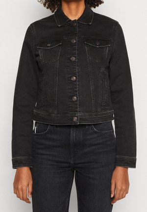 Person iført en tætsiddende sort denimjakke med knaplukning og brystlommer, kombineret med sorte højtaljede jeans.