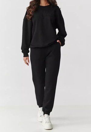 Frau trägt schwarzen langärmligen Sweatshirt und passende schwarze Jogginghose mit weißen Sneakers, läuft vor schlichtem hellem Hintergrund.