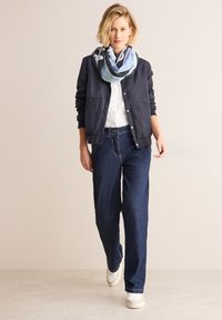 Marineblå bomberjakke med knaplukning, hvid skjorte, mørkeblå brede jeans, cremegule slip-on sko og et mønstret multicolor tørklæde.