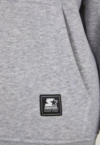 Grå sweatshirtstof med en blød tekstur. Har et sort firkantet logo-patch med hvid tekst og et stjernedesign; detaljer om den forreste lomme er synlige.