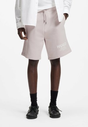 Homme portant un short HUGO rose clair avec cordon, des chaussettes noires et des mocassins noirs, une main dans la poche et une chemise blanche.