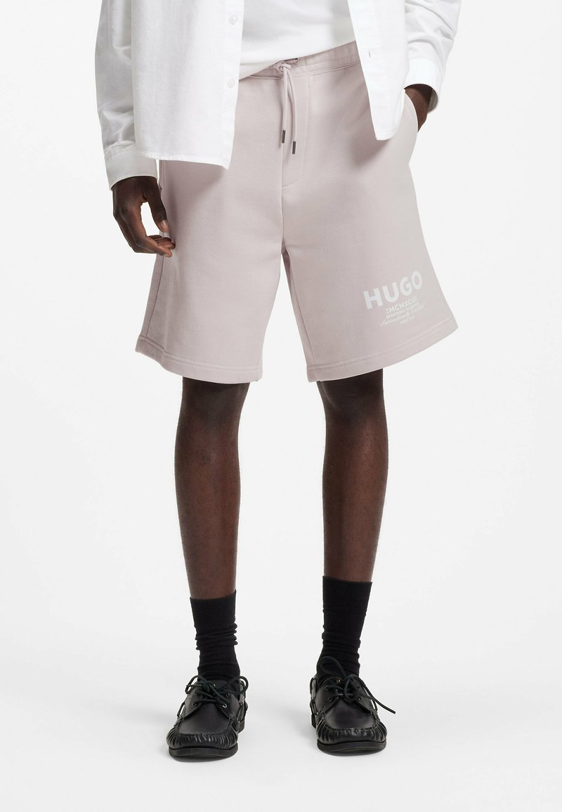 Mann trägt hellrosa HUGO-Shorts mit Kordelzug, schwarze Socken und schwarze Mokassins, mit einer Hand in der Tasche und weißem Hemd.