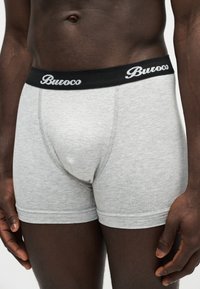 Gros plan d’un homme portant un boxer gris clair avec une ceinture noire portant l’inscription « Buroco ».