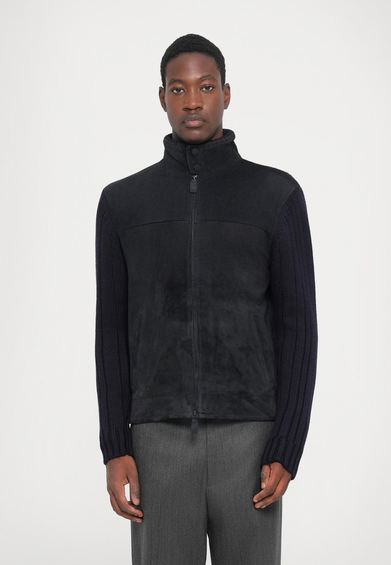 Gilet en polaire noir avec un col montant et une fermeture éclair, manches en tricot bleu marine côtelé et surface texturée. Un pantalon gris complète le look.