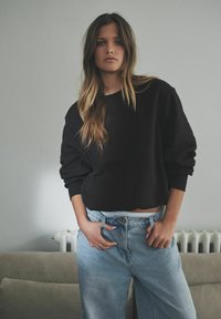 Czarny skrócony sweatshirt z zaokrąglonym dołem, zestawiony z jasnoniebieskimi dżinsami o wysokim stanie. Naturalne światło tworzy subtelne cienie.