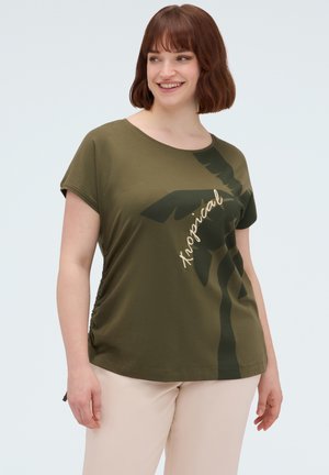 Kvinde smilende, iført en olivengrøn t-shirt med mørkt palmemotiv og teksten "Tropical", kombineret med lyse beige bukser.
