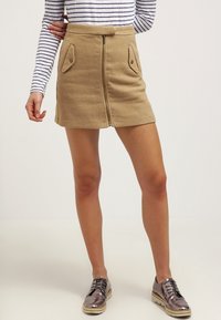 Jupe mini beige texturée avec une fermeture éclair sur le devant et deux poches inclinées, assortie à des chaussures à lacets métalliques et un haut rayé.
