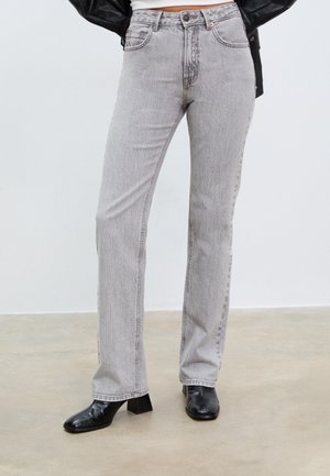 Sive kavbojke ravnega kroja, visoke pasu, pet žepov in subtilne teksture, v kombinaciji z črnimi čevlji na peto do gležnja.