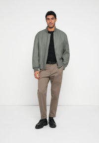 Chaqueta bomber de cuero gris sobre una camiseta negra, combinada con pantalones beige y zapatos negros, sobre un fondo blanco liso.