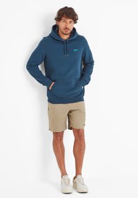 Blue hoodie with a front kangaroo pocket, drawstring hood, and logo patch. Paired with beige shorts and light sneakers. Smooth texture.